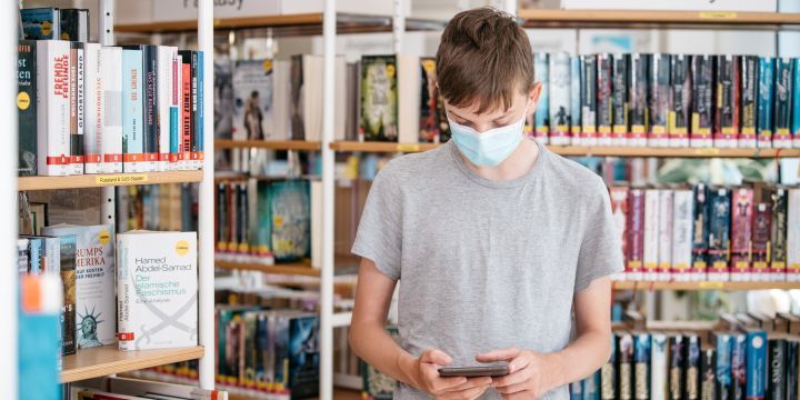 Junge steht mit Smartphone zwischen Bücherregalen in der Stadtteilbibliothek Lobeda  ©JenaKultur | C. Worsch