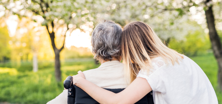 Eine blonde Frau umarmt eine ältere Person im Rollstuhl man sieht sie von hinten