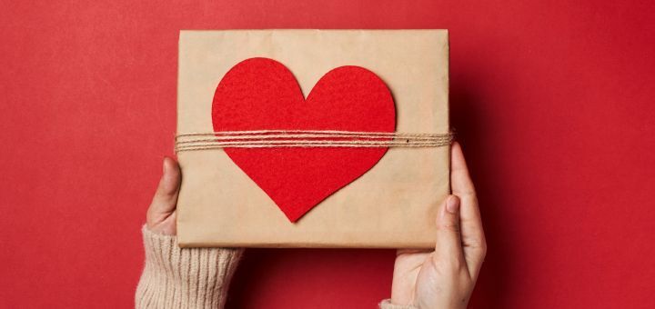 Zwei Hände halten ein braunes Geschenk mit einem roten Herzen darauf über einen roten Hintergrund