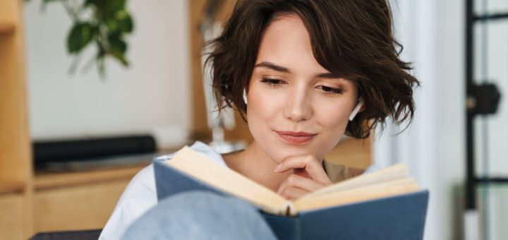 Eine Frau mit kurzen braunen Haaren hat Kopfhörer auf und liest ein Buch 