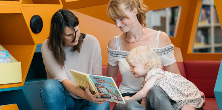 Bibliothekarin liest Kind vor, das bei Mama auf dem Schoß sitzt