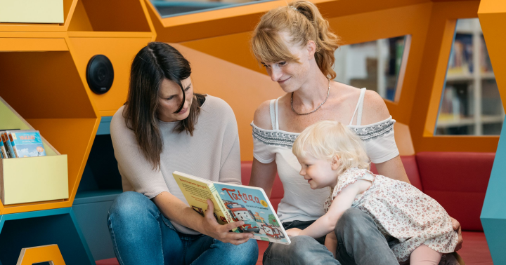 Bibliothekarin liest Kind vor, das bei Mama auf dem Schoß sitzt